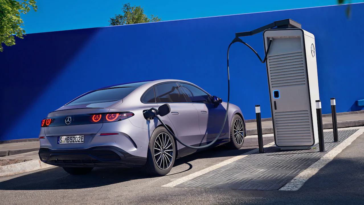 Auto elettrica Mercedes-Benz di colore grigio opaco in fase di ricarica presso una colonnina di design ultra-rapida su sfondo blu.